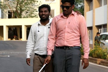 Photo of two men walking along a street. One man has a white cane. The other touches him at the shoulder and let hims guide him. 