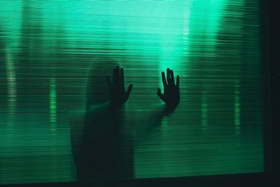 Photo of a woman holding her hands against a green glass surface with slats. Through the structure of the glass surface you can see only the silhouette of the woman.