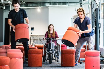 Photo of two men moving stools aside so that a woman in a wheelchair can pass.