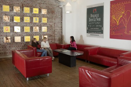 Photo of the lobby at the Dialoghaus Hamburg: a group of red soafas with some visitors seated on them; on the walls portraits of the guides and posters of other dialog venues.