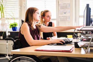 Photo of a woman in a wheelchair, who is explaining something to a co-worker sitting next to her, on a computer