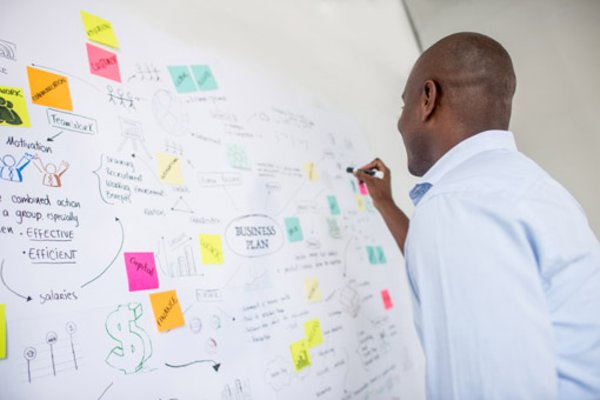 Photo of a man writing something on a white board which is covered with post-its and sketches.
