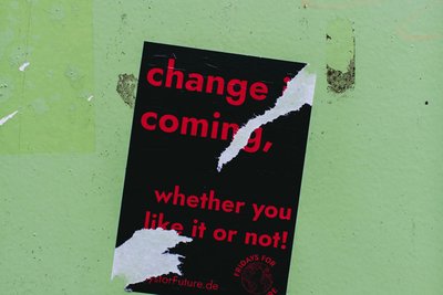 Photo of a sticker on a green wall, half torn off, saying "Change is coming - whether you like it or not"