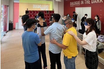 Photo of participants of a workshop. The paricipants wear blindfolds and queue up, holding the person in front by the shoulder.