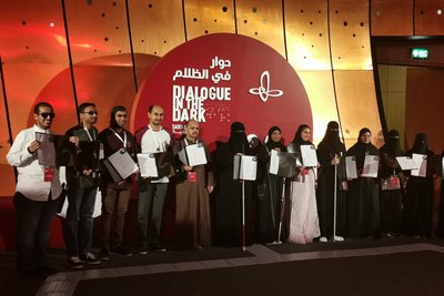 Photo of the dialogue in the DArk team in Dhahran, Saudi Arabia, at the certification ceremony. The team mebmer stand infront of the Dialogue in the Dark logo, proudly presenting their certificates for the passed training as guides in the dark.