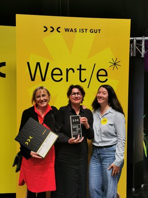 Drei Frauen stehen vor dem gelben Logo "was ist gut" mit ihrer Auszeichnung