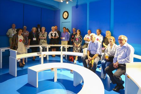 A photo of a vistor group in the Dialogue with Time room "Future of Aging", where the visitors are seated on circular benches, watching a video on enviromental changes needed to meet an ageing society. 