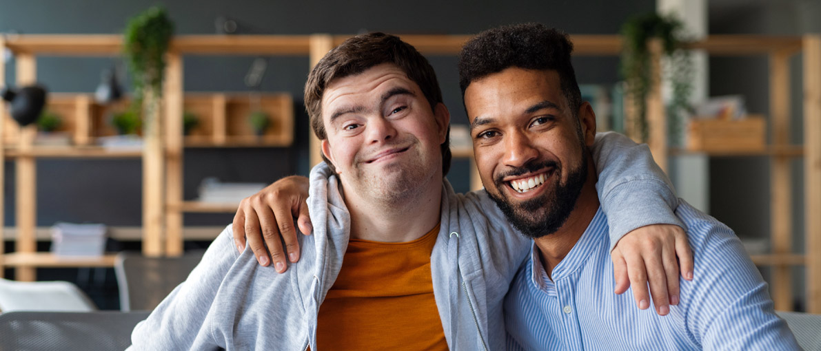 Foto von einem jungen Mann mit Down Syndrom und einem jungen Schwarzen Mann, die sich gegenseitig den Arm um die Schulter gelegt haben und in die Kamera lächeln.